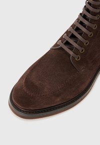 Stivale in suede marrone con punta arrotondata, cuciture in vista, occhielli metallici e lacci marroni, dotato di una suola in pelle marrone e superficie testurizzata.