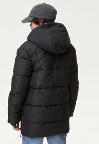 Giacca a vento nera con cuciture verticali, cappuccio staccabile, zip frontale e due tasche laterali, abbinata a un berretto grigio e jeans azzurri chiari.