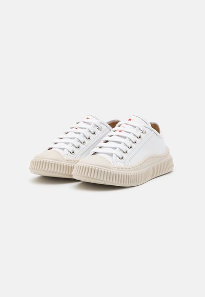 Sneakers bianchi in tela con suole in gomma beige, punta rotonda e occhielli in metallo argentato. I lacci sono bianchi con dettagli rossi sulla linguetta.