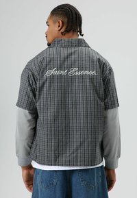 Kurzärmliges Hemd mit Karomuster in Blau-, Grau- und Weißtönen, hinten mit dem Logo "Saint Essence" in Kursivschrift. Darüber ein grau langärmliges Shirt getragen.