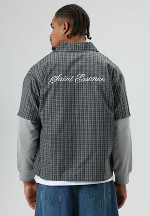 Geruit overhemd met korte mouwen in blauw, grijs en wit, met op de rug een logo "Saint Essence" in cursief. Draagt een grijze longsleeve eronder.