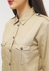 Camicia beige a bottoni con colletto, due tasche sul petto e dettagli neri sui bottoni; realizzata in tessuto liscio e leggero.
