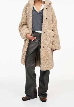Personne portant un long manteau beige duveteux, une chemise grise boutonnée sur un haut blanc, un pantalon noir à jambes larges et des chaussures marron, debout avec la main dans la poche.