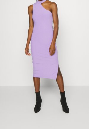 Kobieta ubrana w bezrękawnikową, liliową, prążkowaną sukienkę midi z asymetrycznym dekoltem i czarnymi botkami, stojąca na jednolitym tle.