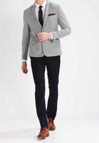 Man justerar en grå blazer med en bourgogne fickduk, bär svart byxor, vit skjorta, svart slips och bruna dressade skor, går framåt.