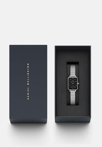 Montre carrée noire avec un bracelet en argent texturé, présentée dans une boîte bleu marine. La boîte dispose d'un texte doré et d'un intérieur noir doux.