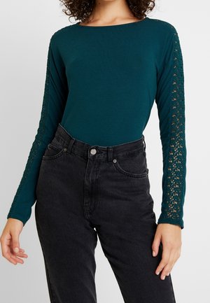 Femme portant un haut à manches longues couleur sarcelle avec des détails en dentelle sur les manches et un jean noir taille haute sur un fond uni.