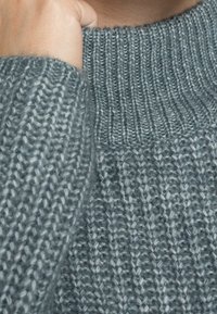 Pull en maille épaisse gris clair avec un col montant côtelé. Présente un motif texturé avec des points variés pour plus de profondeur.