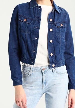 Personne portant une veste en jean bleu foncé sur une chemise blanche et un jean bleu clair, debout avec une main visible sur le côté.