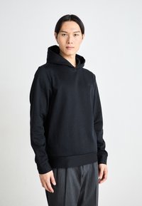 Sweatshirt à capuche noir en tissu texturé avec des rayures verticales. Il comporte une poche kangourou à l'avant et des poignets et un ourlet côtelés.