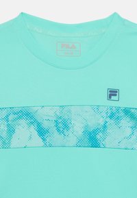 Chemise turquoise clair à encolure ronde, avec un motif texturé dans des teintes plus sombres de bleu. Logo Fila en haut à gauche.