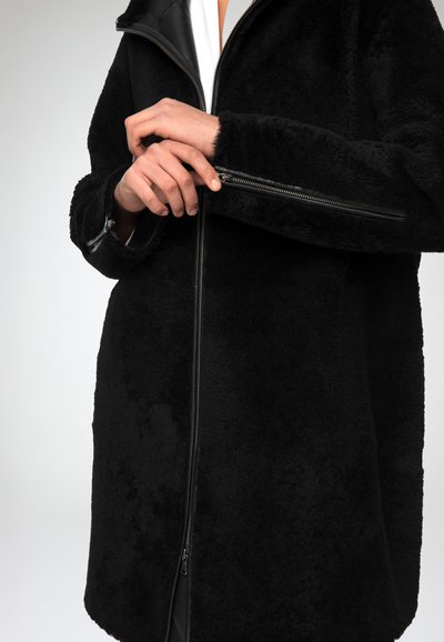 Cappotto in pile nero con zip anteriore, tessuto testurizzato e colletto in pelle staccabile. Primo piano su mani che regolano la zip.