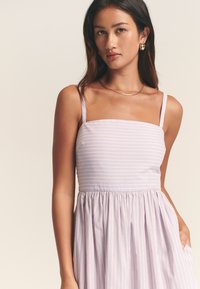 Lilakų dryžuota suknelė su kvadrato formos iškirpte, plonomis petnešėlėmis ir susegta sijono dalimi. Pagaminta iš minkštos medžiagos, turi šonines kišenes. Papuošta metalinėmis auskarais.