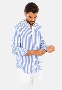 Camicia di lino a righe blu e bianche con colletto alla mandarina, chiusura con bottoni e maniche arrotolate, indossata con pantaloni bianchi.