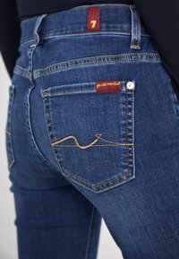 Dark blue denim jeans with a close-fitting design. Features a red label with the number “7” and a signature pocket design with gold stitching.