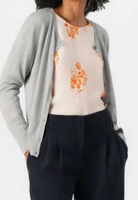 Světle šedý pletený cardigan s dlouhými rukávy, s knoflíkovým zapínáním a žebrovanými manžetami, nošený přes broskvovou blůzu s oranžovým květinovým potiskem.
