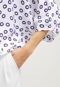 Bras partiellement inséré dans la poche d'un pantalon blanc, portant un chemisier blanc avec un motif circulaire bleu et des manches bouffantes.