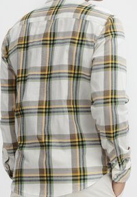 Mann in einem Langarm-Hemd mit kariertem Muster in Gelb, Grün, Braun und Grau, mit einer Hand in der Tasche seiner beigen Hose.