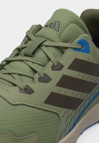 Κοντινή λήψη πράσινου αθλητικού παπουτσιού με ύφασμα από δίχτυ, κλείσιμο με κορδόνια στο μπροστινό μέρος, σκούρες ρίγες και μπλε λεπτομέρεια κοντά στην περιοχή του αστραγάλου.