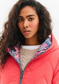 Korálová prešívaná bunda s kvetinovou vnútornou podšívkou, vybavená vysokým golierom a zeleným zipsom. Nosená cez vrstvený béžový sveter s textúrou.