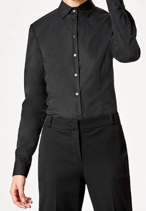 Personne portant une chemise noire cintrée à manches longues boutonnée, rentrée dans un pantalon noir taille haute sur fond blanc.
