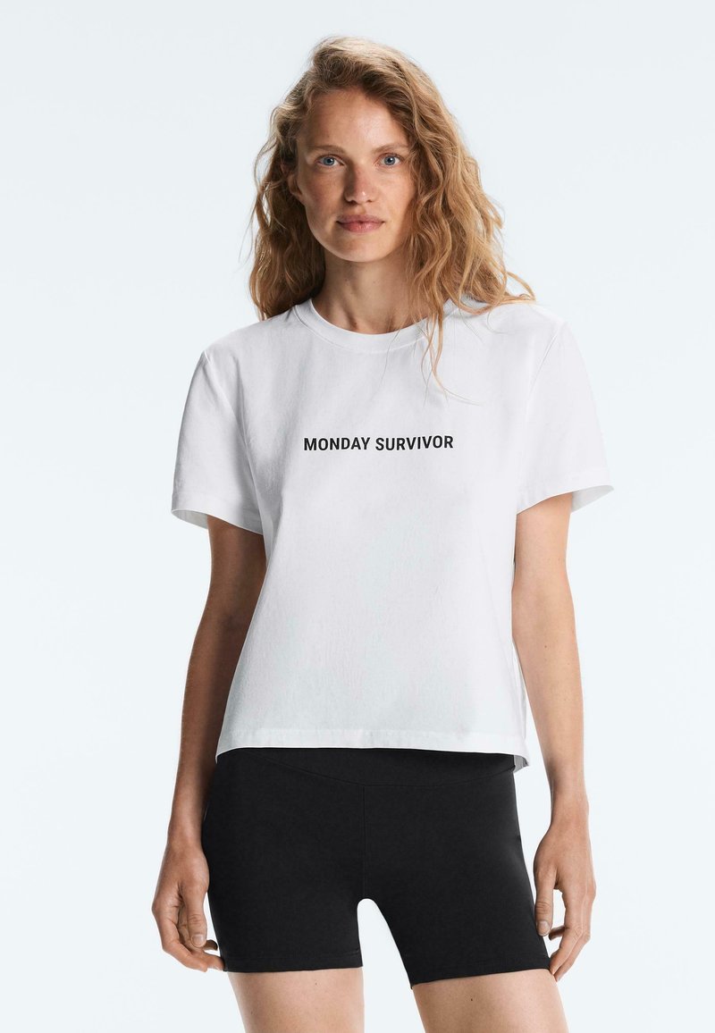 Kvinde med bølget blondt hår iført en hvid T-shirt med teksten "MANDAGSOVERLEVER" og sorte shorts, stående foran en enkel lys baggrund.