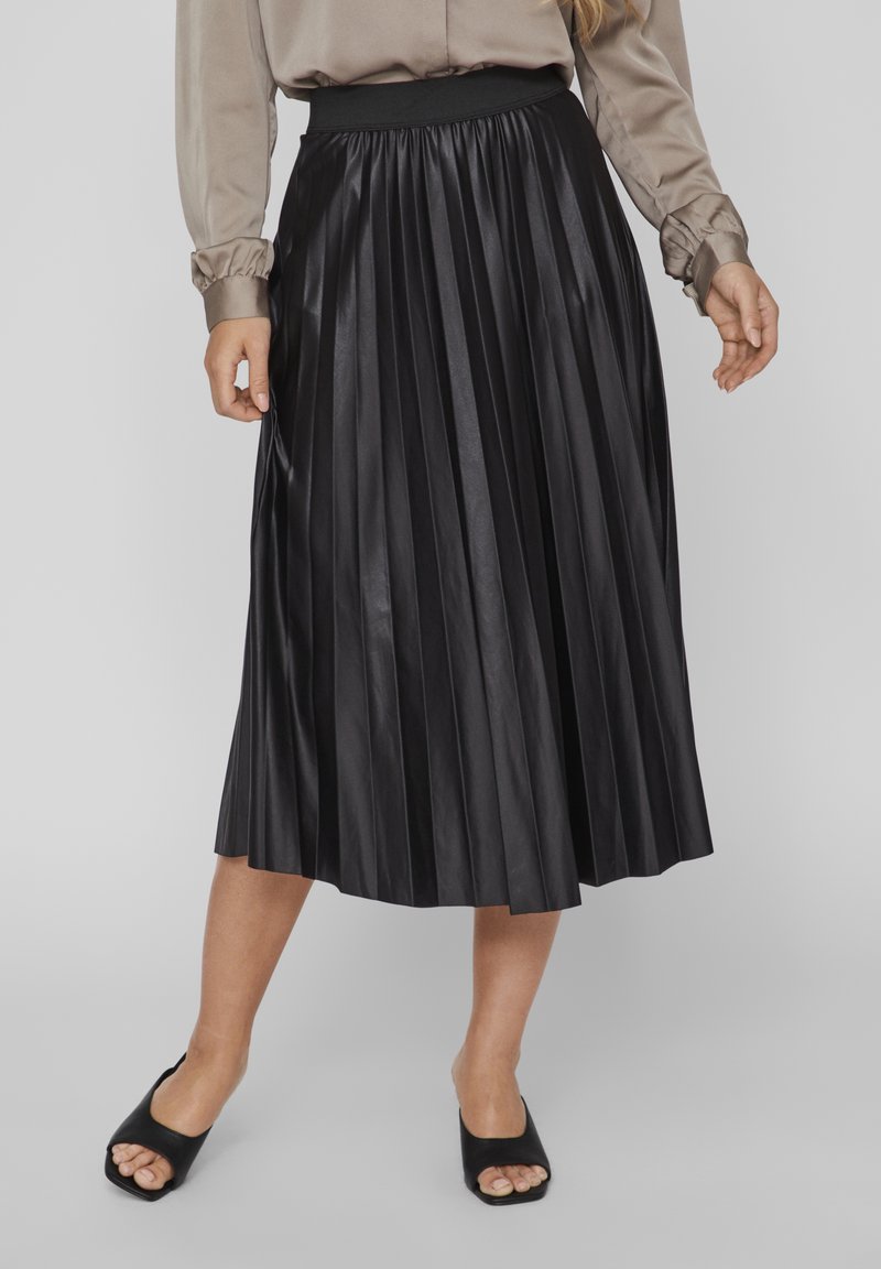 Femme portant une jupe midi plissée noire, un chemisier beige à manches longues et des mules à talons noires à bout ouvert sur un fond gris clair.