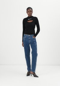 Top nero a maniche lunghe con un taglio, abbinato a jeans blu a vita alta con borchie laterali. Scarpe con tacco nere completano l'outfit.