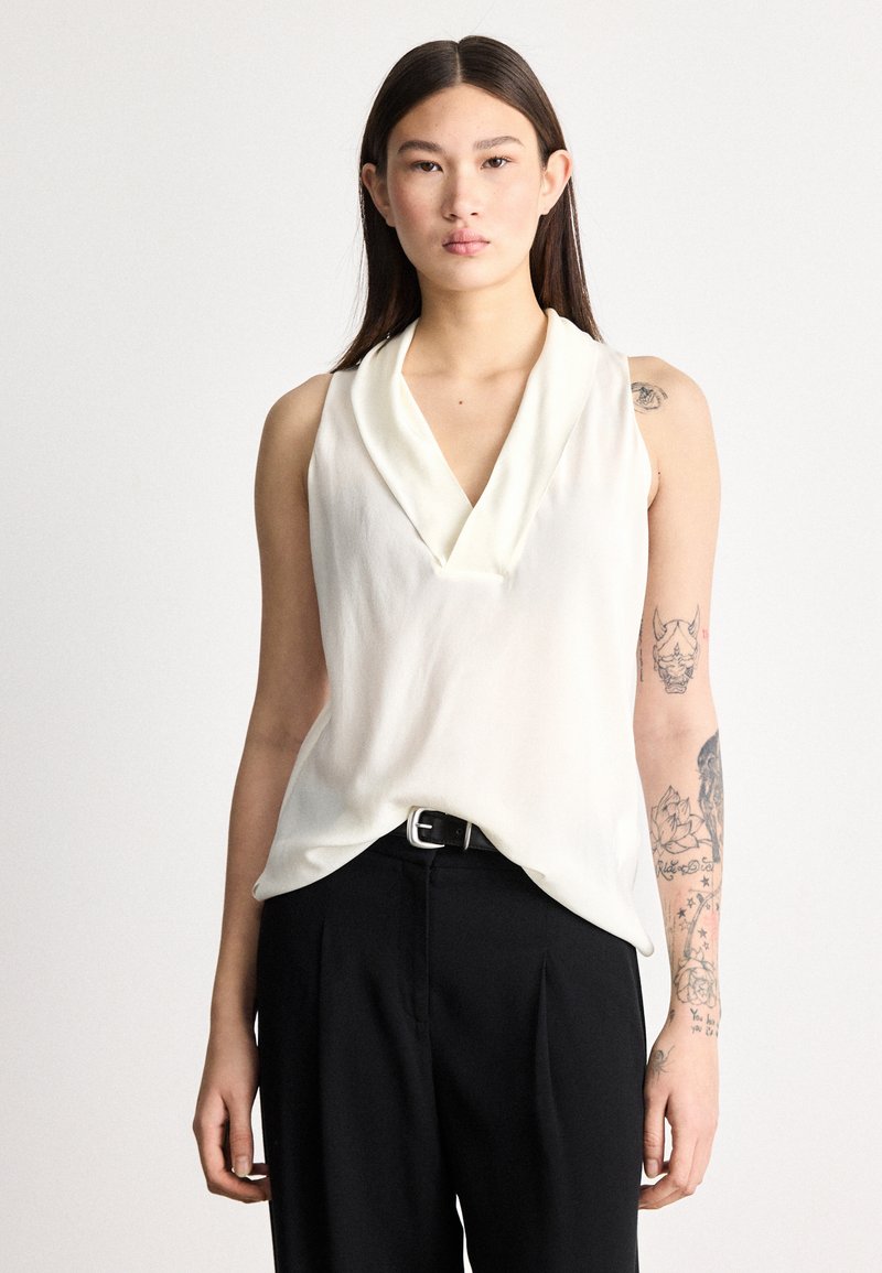 Αμάνικη μπλούζα από εκρού ύφασμα με ντραπαρισμένο ντεκολτέ σε σχήμα V. Συνδυασμένη με μαύρο, ραμμένο παντελόνι που διαθέτει ασημένια αγκράφα ζώνης.