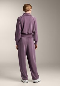 Lila Trainingsanzug mit entspannter Passform, mit Kragen, langen Ärmeln und elastischen Bündchen. Weicher Stoff, kombiniert mit weißen Sneakers.