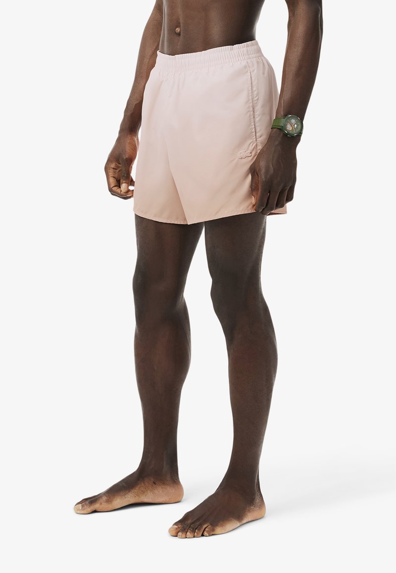 Costume da bagno rosa chiaro realizzato in tessuto morbido, dotato di cintura elastica e un piccolo logo; il modello è in piedi a piedi nudi con le braccia rilassate.