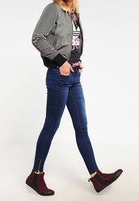 Chaqueta bomber gris, puños acanalados negros, camiseta negra con flores, jeans ajustados azules y botas de tobillo burdeos con acentos negros.