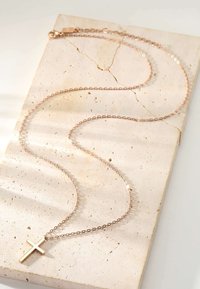 Halskette aus Roségold mit einem schlichten Kreuzanhänger, der auf einer strukturierten, cremefarbenen Steinfäche liegt.
