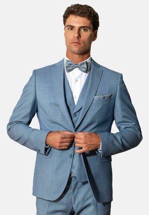 Man wearing light blue tuxedo, white shirt, and matching bow tie, buttoning jacket with neutral expression against plain background.