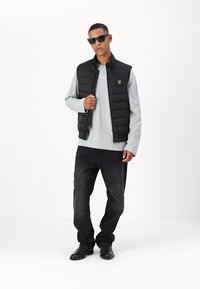 Black puffer vest with zip closure over a light gray long-sleeve shirt, paired with dark jeans and sunglasses. Prominent logo on the vest.