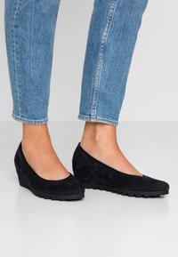Chaussures à talon wedge en daim noir avec un design de bout arrondi, présentant une surface texturée et un petit talon wedge. Associées à un jean bleu clair.