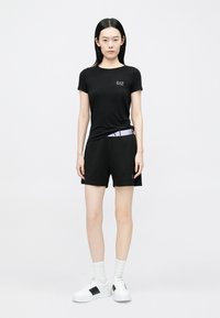 Tricou negru mulat cu logo mic EA7, asortat cu pantaloni scurți negri cu talie violet deschis. Include adidași albi cu accente negre.