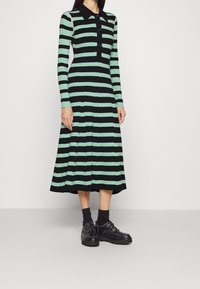 Abito a righe lavorato a maglia in verde e nero con maniche lunghe, collo a camicia, chiusura con bottoni e orlo svasato. Abbinato a scarpe nere e calzini.