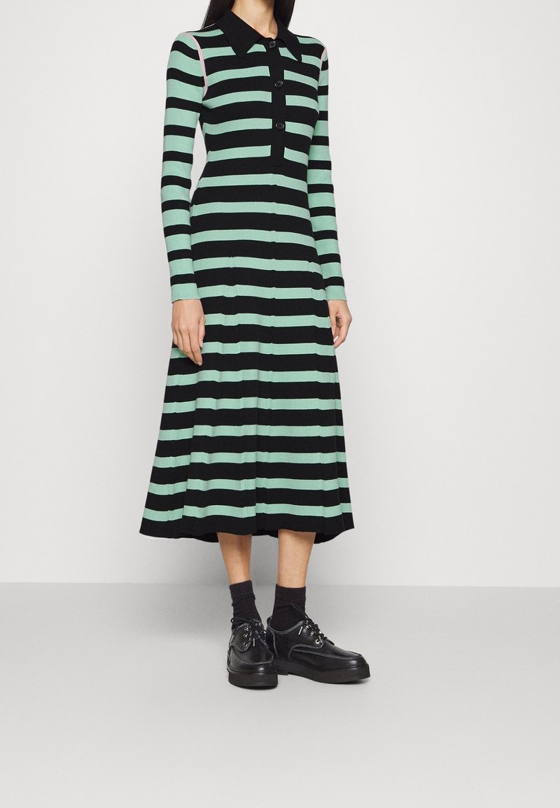 Abito a righe lavorato a maglia in verde e nero con maniche lunghe, collo a camicia, chiusura con bottoni e orlo svasato. Abbinato a scarpe nere e calzini.