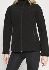 Kvinde iført en sort softshell-jakke med lynlås, lynlåslommer og justerbare håndledsremme.