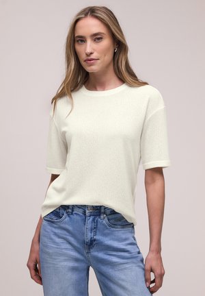 Frau mit langen blonden Haaren, die ein weißes kurzärmeliges Strickoberteil und hellblaue Jeans vor einem schlichten Hintergrund trägt.