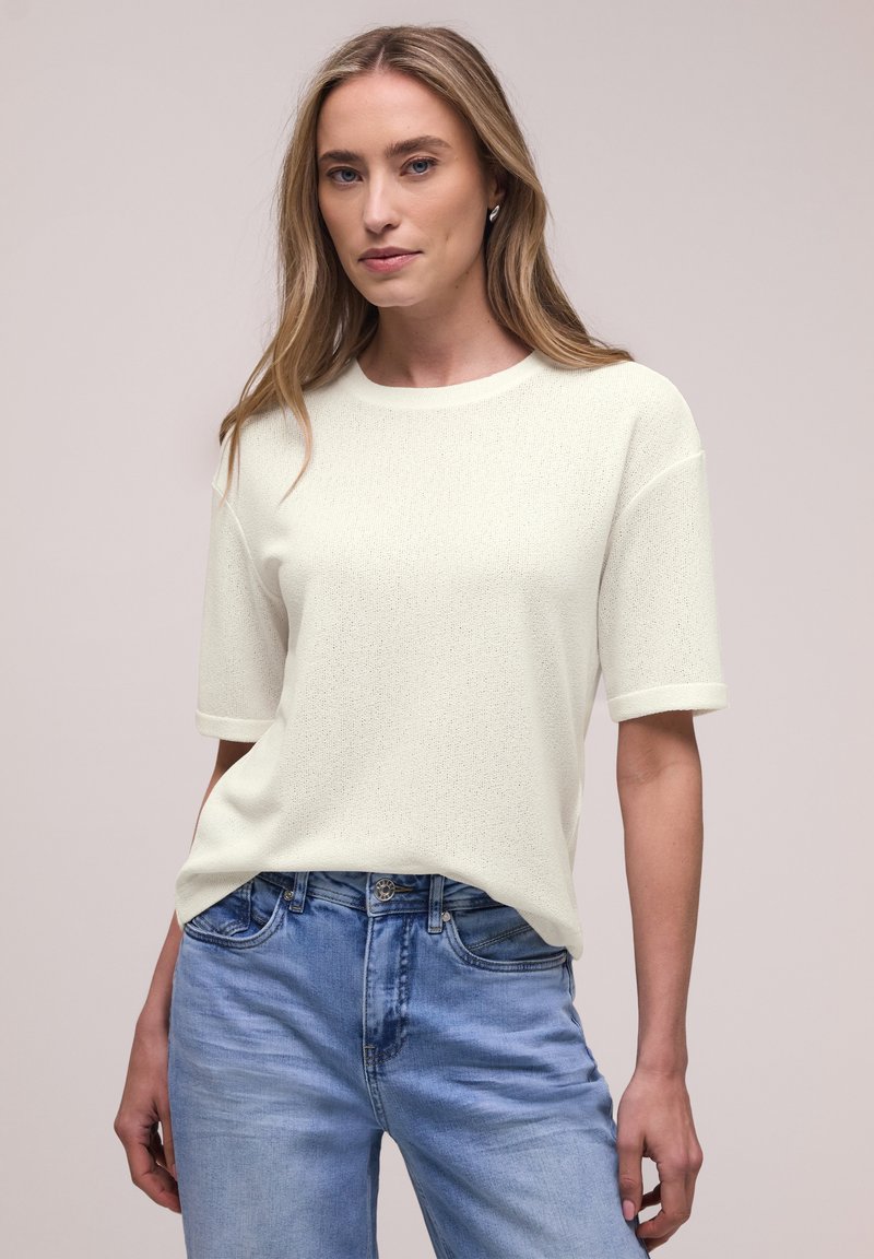 Frau mit langen blonden Haaren, die ein weißes kurzärmeliges Strickoberteil und hellblaue Jeans vor einem schlichten Hintergrund trägt.