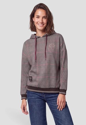 Mujer sonriendo con una sudadera con capucha de cuadros con cordones burdeos y vaqueros azules, de pie frente a un fondo liso y claro.