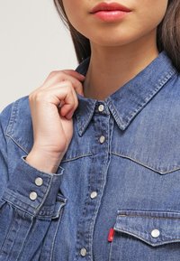 Chemise en denim avec un col pointu, des boutons-pression argentés et une poche sur la poitrine. Couleur bleu clair avec des détails de couture visibles.