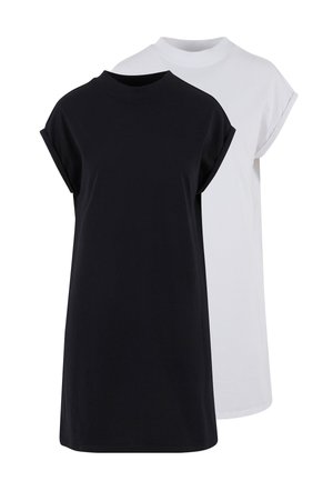 Femme aux cheveux bouclés portant une robe noire simple à manches courtes, debout devant un fond gris clair uni.