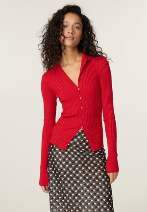 Vrouw met krullend haar, die een rood geribd gebreid vest met knopen en een zwarte rok met een wit hartjespatroon draagt, staat voor een effen achtergrond.