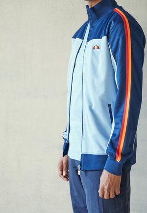 Personne portant une veste de sport bleue avec fermeture éclair, avec des bandes rouges et orange sur les manches et un petit logo sur la poitrine, debout contre un mur uni.