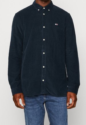 Homme portant une chemise en velours côtelé bleu foncé à boutons avec un petit logo sur la poitrine et un jean bleu, debout devant un fond uni.