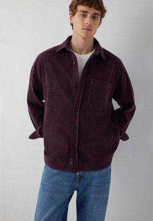 Young man wearing a dark purple corduroy button-up shirt over a white t-shirt and blue jeans, standing with hands behind his back.