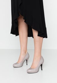 Tacones altos plateados brillantes con punta redondeada y delgado tacón de aguja negro, con un acabado liso de purpurina y silueta curvada.